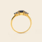 A gold ring featuring a central dark blue oval gemstone and three smaller blue oval gemstones on either side, set against a solid light beige background.