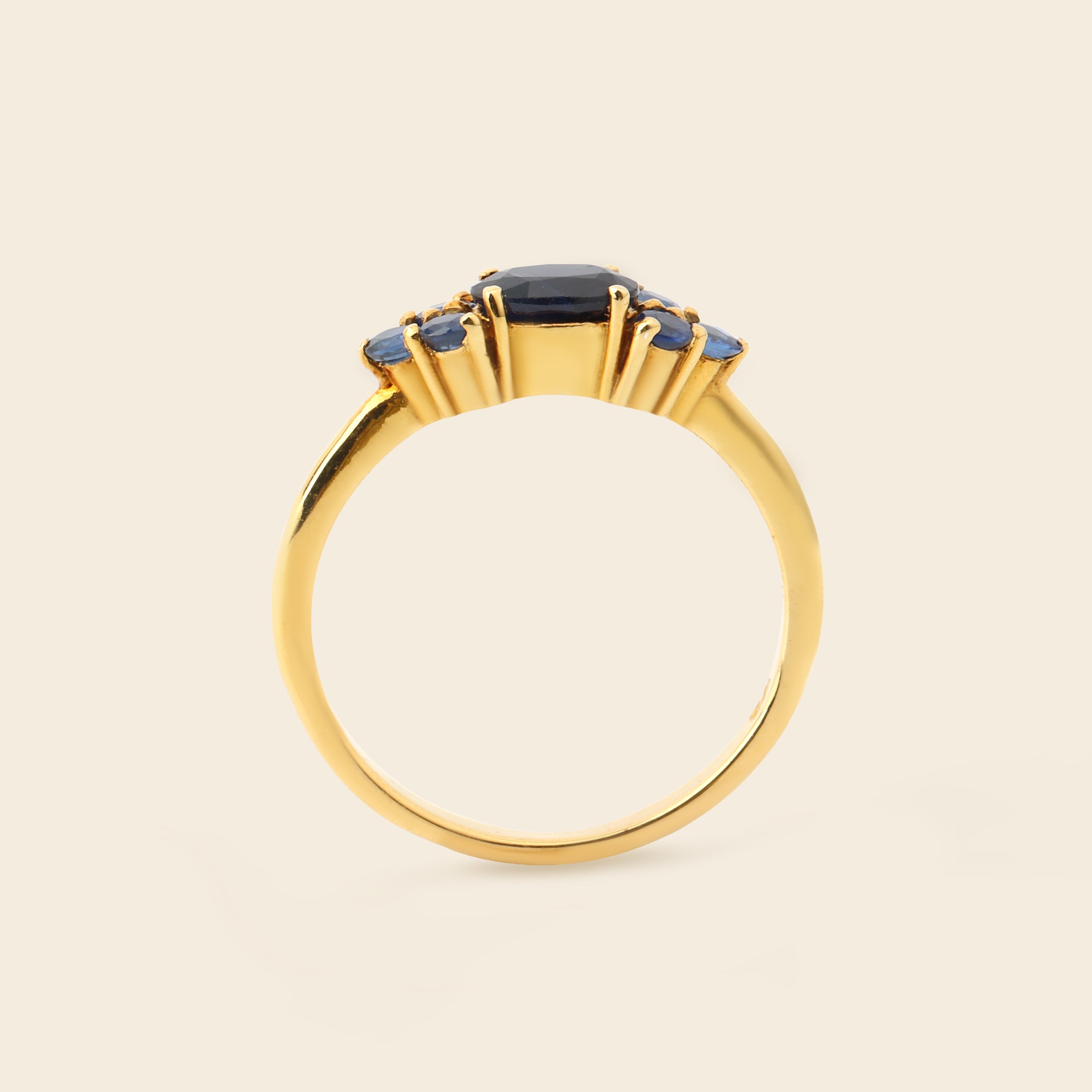 A gold ring featuring a central dark blue oval gemstone and three smaller blue oval gemstones on either side, set against a solid light beige background.