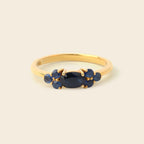 A gold ring featuring a central oval dark blue gemstone flanked by two clusters of small round blue gemstones, set against a plain light beige background.