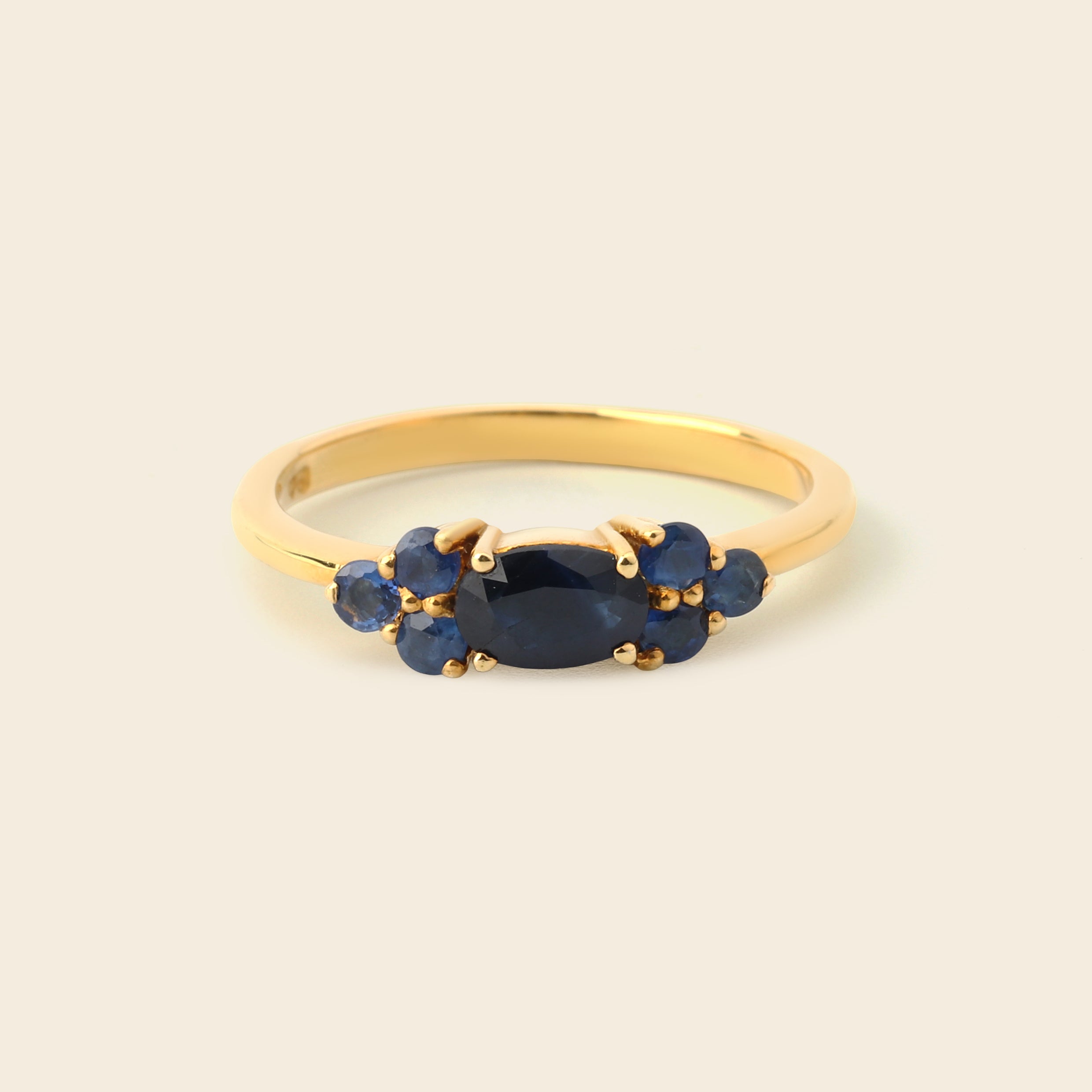A gold ring featuring a central oval dark blue gemstone flanked by two clusters of small round blue gemstones, set against a plain light beige background.