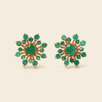 A pair of gold starburst stud earrings with green gemstones, displayed on a light beige background.