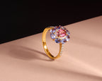 A gold ring with a central oval pink gemstone surrounded by a halo of round purple and blue gemstones, accented with small clear stones on the band, set against a beige and black background.