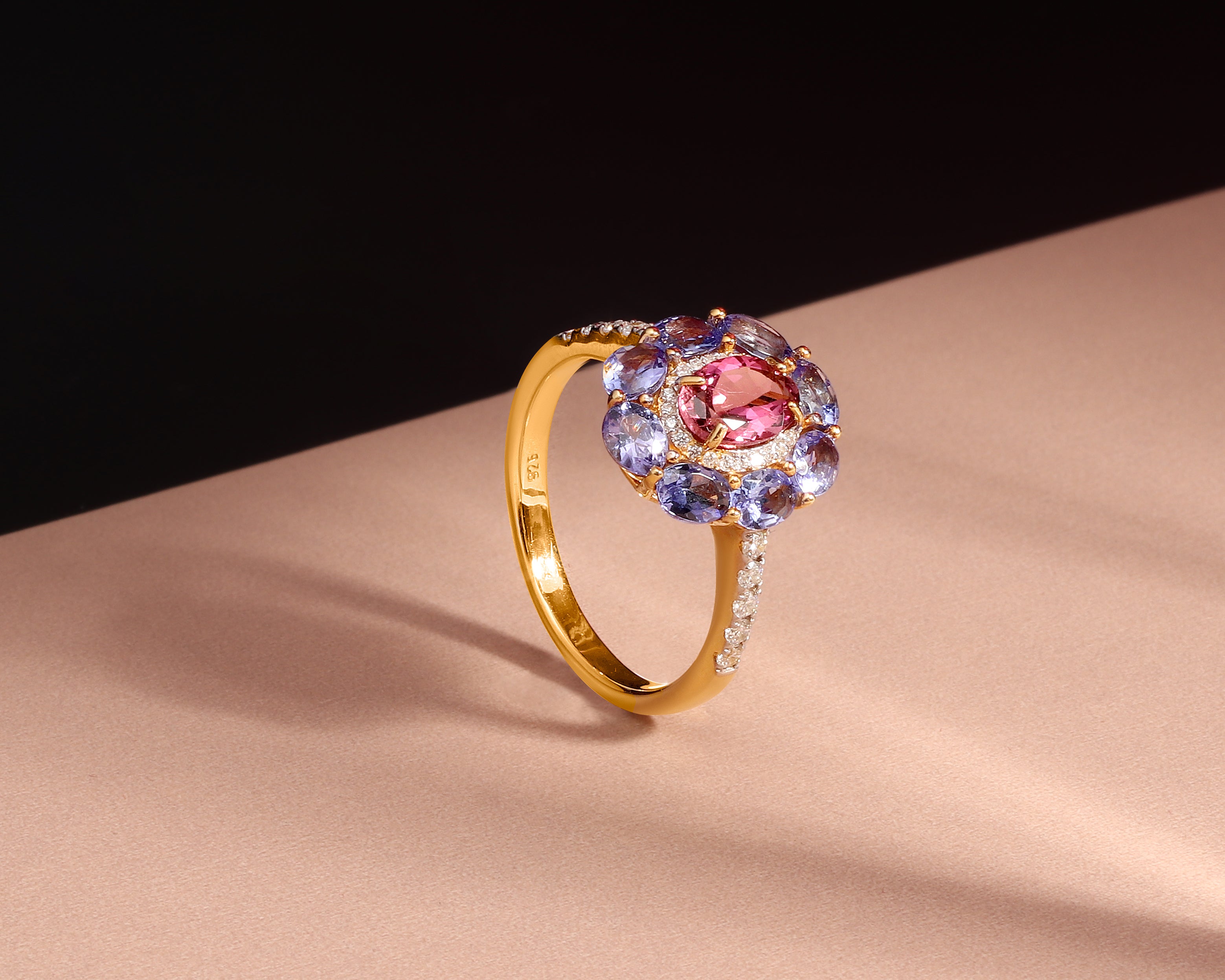 A gold ring with a central oval pink gemstone surrounded by a halo of round purple and blue gemstones, accented with small clear stones on the band, set against a beige and black background.
