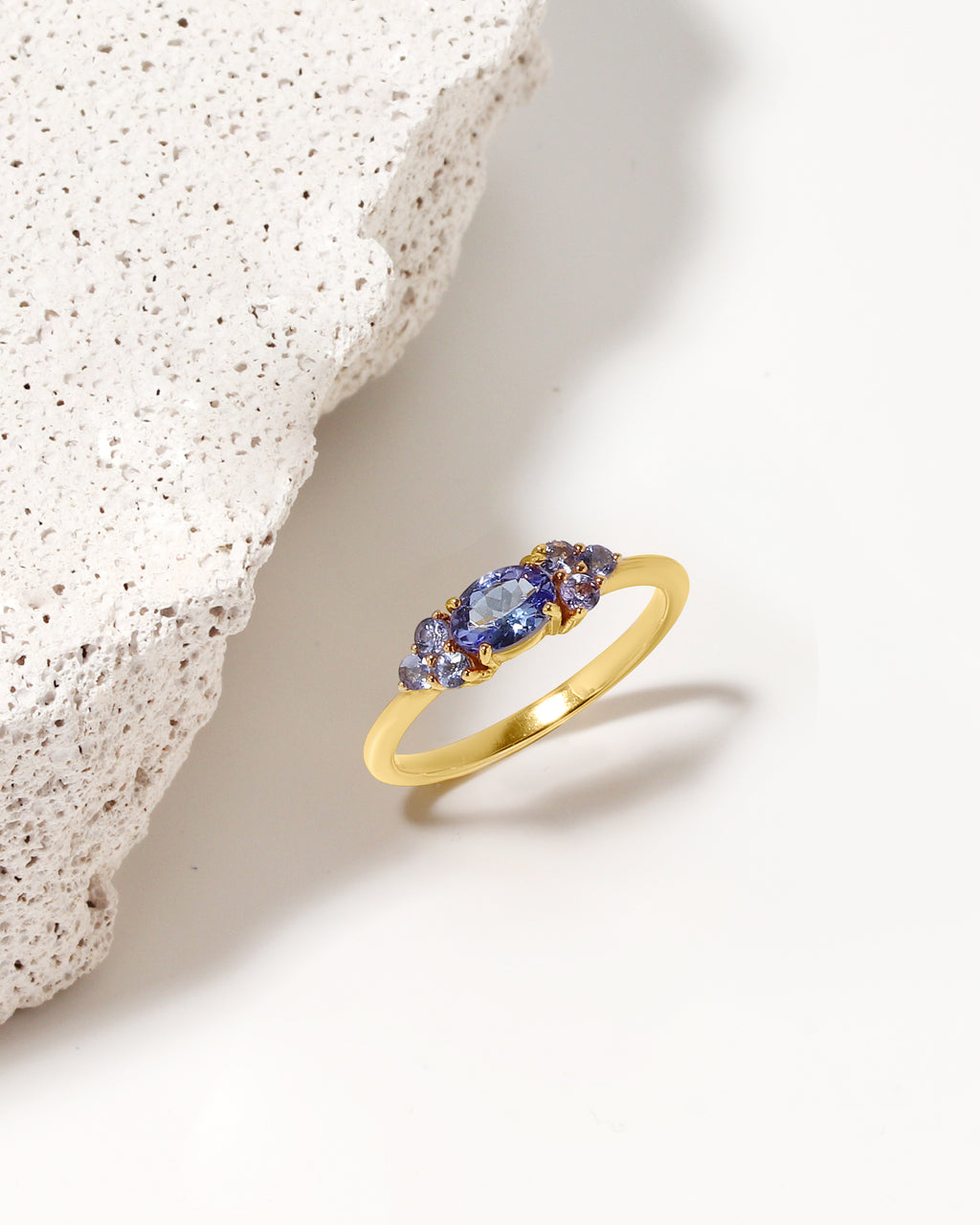 A gold ring with three marquise-cut blue gemstones, styled on a clean white surface next to a textured white stone prop.