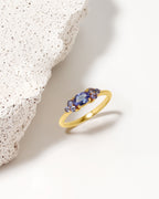 A gold ring with three marquise-cut blue gemstones, styled on a clean white surface next to a textured white stone prop.