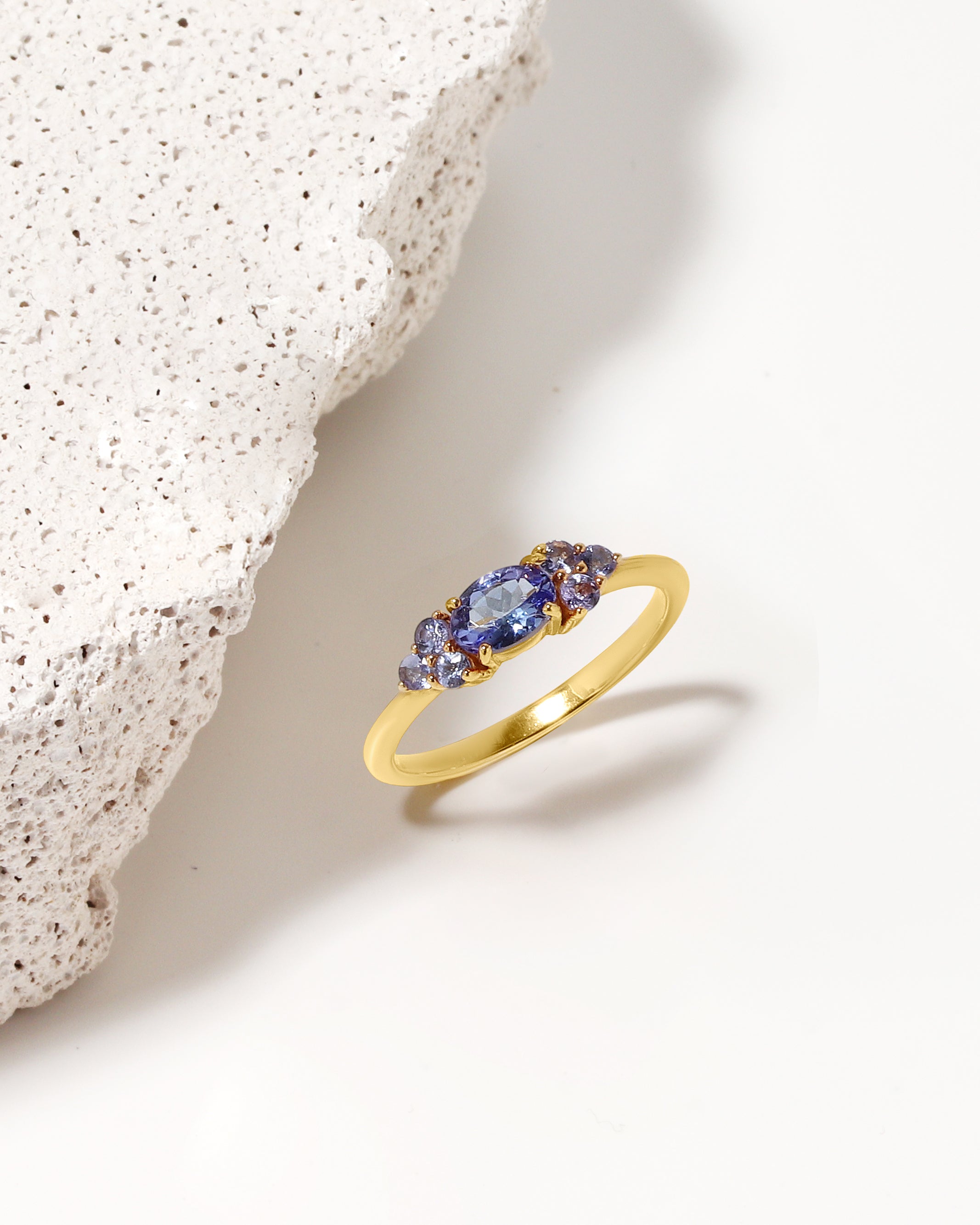 A gold ring with three marquise-cut blue gemstones, styled on a clean white surface next to a textured white stone prop.