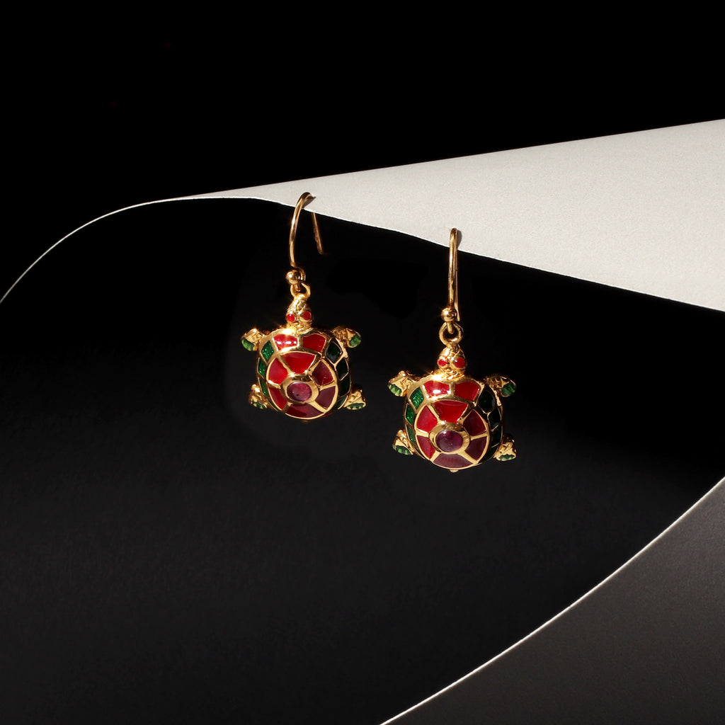 A pair of gold turtle drop earrings with red and green enamel detailing, displayed on a black and white curved surface.