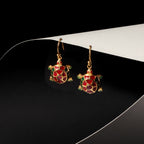 A pair of gold turtle drop earrings with red and green enamel detailing, displayed on a black and white curved surface.
