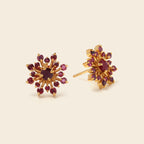 A pair of gold starburst stud earrings featuring a central round dark red gemstone surrounded by smaller round red gemstones, displayed on a light beige background.