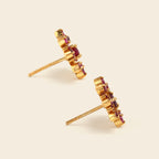 A pair of gold post earrings with a cluster of small round-cut red gemstones, shown on a light beige background.