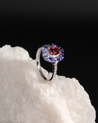 A silver ring featuring a central round red gemstone surrounded by a halo of blue and purple round stones, displayed on a textured white rock against a black background.