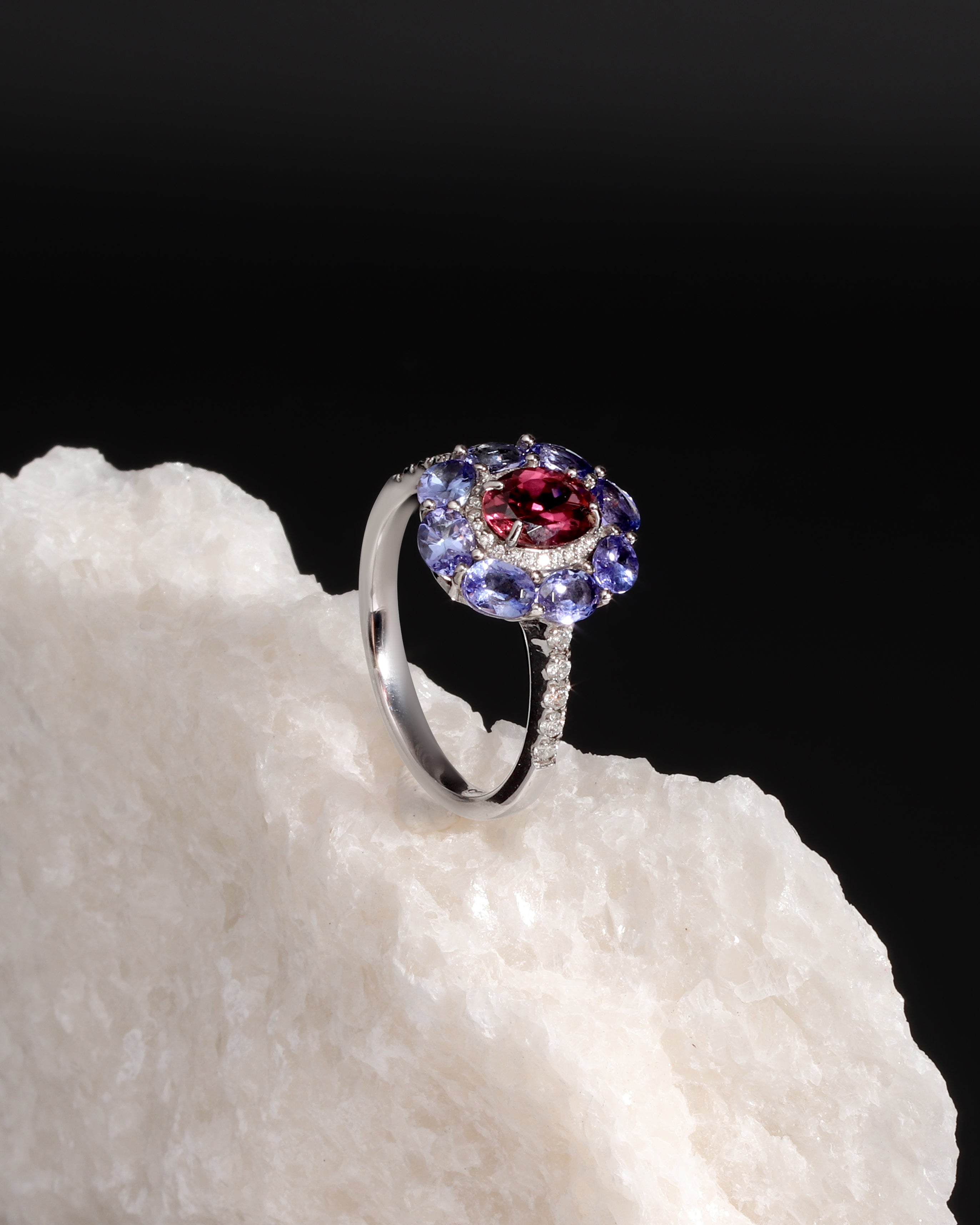 A silver ring featuring a central round red gemstone surrounded by a halo of blue and purple round stones, displayed on a textured white rock against a black background.