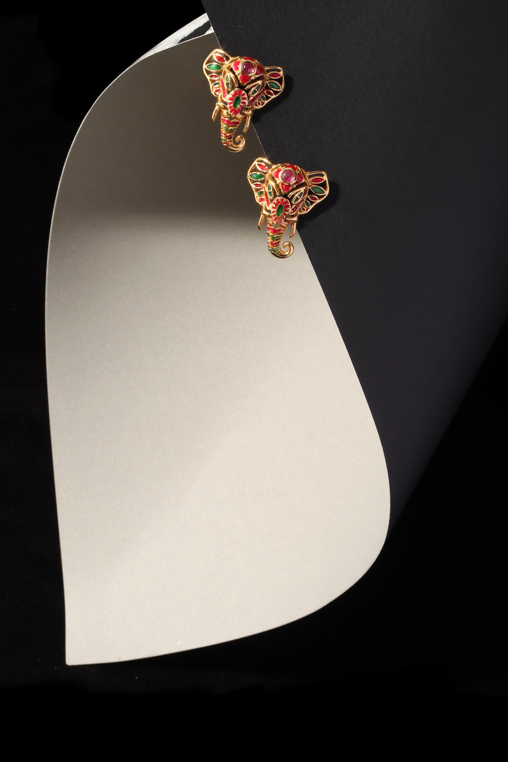 A pair of gold-toned elephant stud earrings with red and green enamel detailing, displayed on a curved white card against a black background.