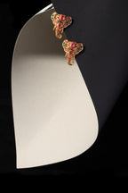 A pair of gold-toned elephant stud earrings with red and green enamel detailing, displayed on a curved white card against a black background.