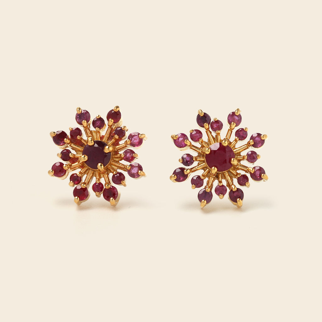 A pair of gold starburst stud earrings set with deep red gemstones, displayed on a light beige background.