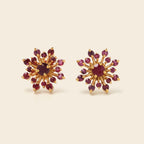 A pair of gold starburst stud earrings set with deep red gemstones, displayed on a light beige background.