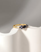 A gold ring with a central dark blue oval stone and small blue round stones on either side, displayed on a white curved stand against a gray background.