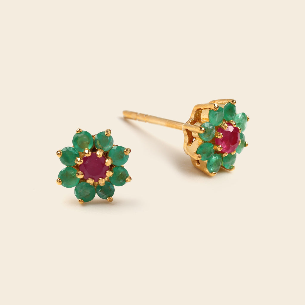 A pair of gold flower-shaped stud earrings featuring green emerald-cut gemstones around a central round pinkish-red ruby center, displayed on a light beige background.