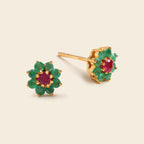 A pair of gold flower-shaped stud earrings featuring green emerald-cut gemstones around a central round pinkish-red ruby center, displayed on a light beige background.