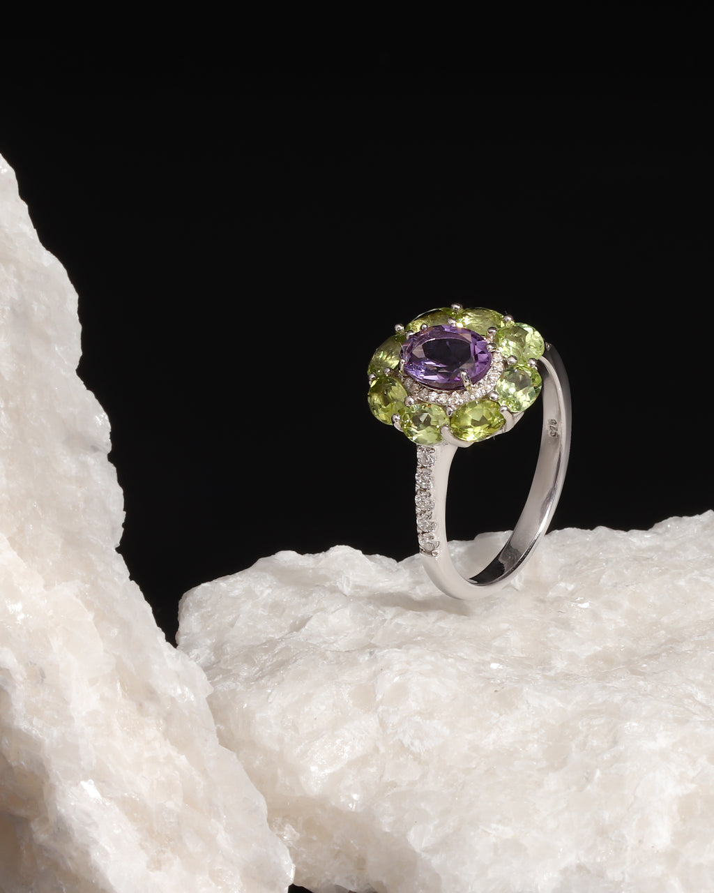 A silver ring featuring a round purple amethyst center stone surrounded by a halo of green peridot and small clear accent stones, displayed on a white textured rock against a solid black background.