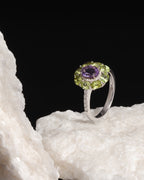 A silver ring featuring a round purple amethyst center stone surrounded by a halo of green peridot and small clear accent stones, displayed on a white textured rock against a solid black background.