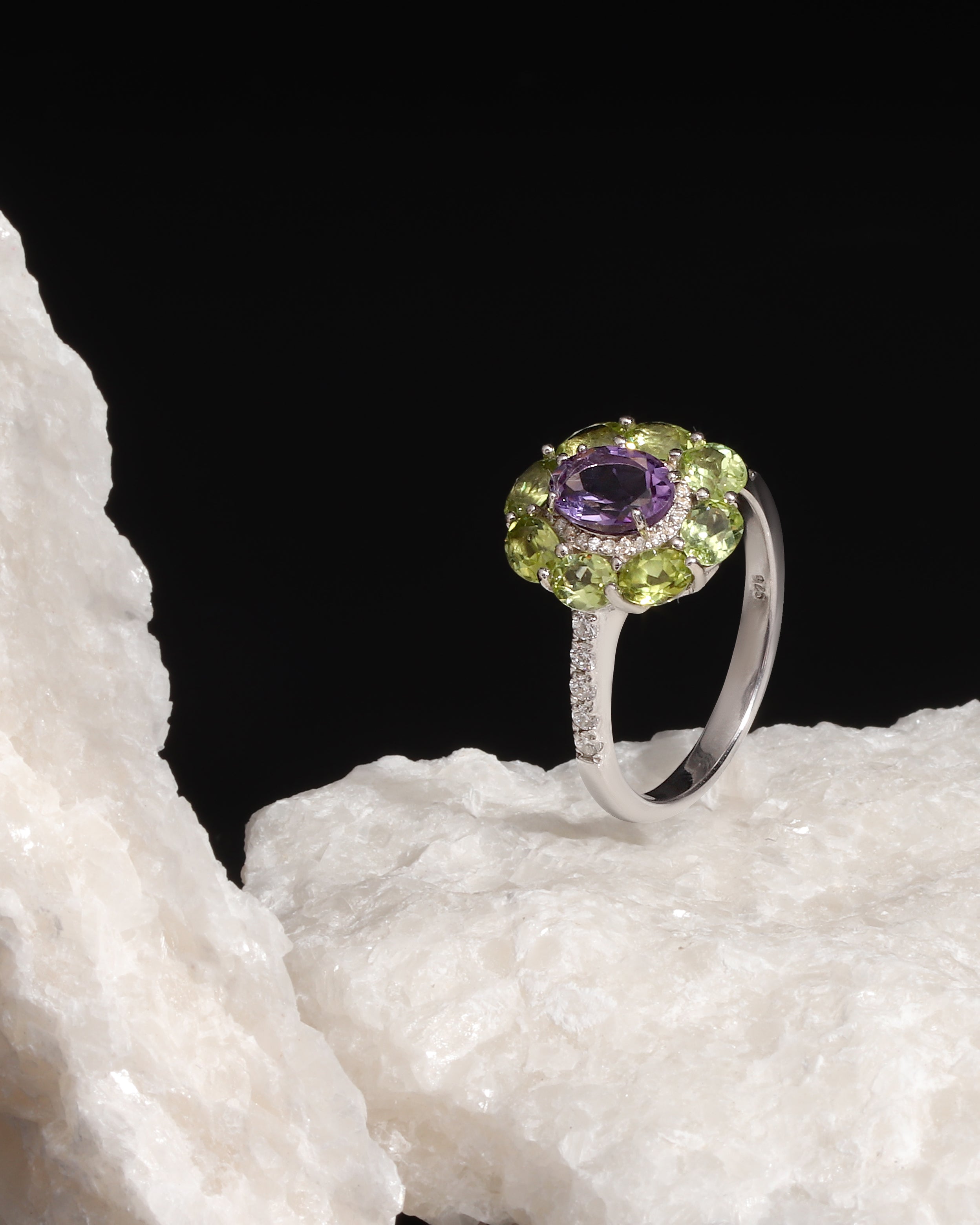 A silver ring featuring a round purple amethyst center stone surrounded by a halo of green peridot and small clear accent stones, displayed on a white textured rock against a solid black background.