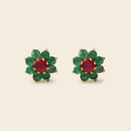 A pair of gold flower-shaped stud earrings featuring a round red center stone surrounded by six green round stones, set against a solid light beige background.