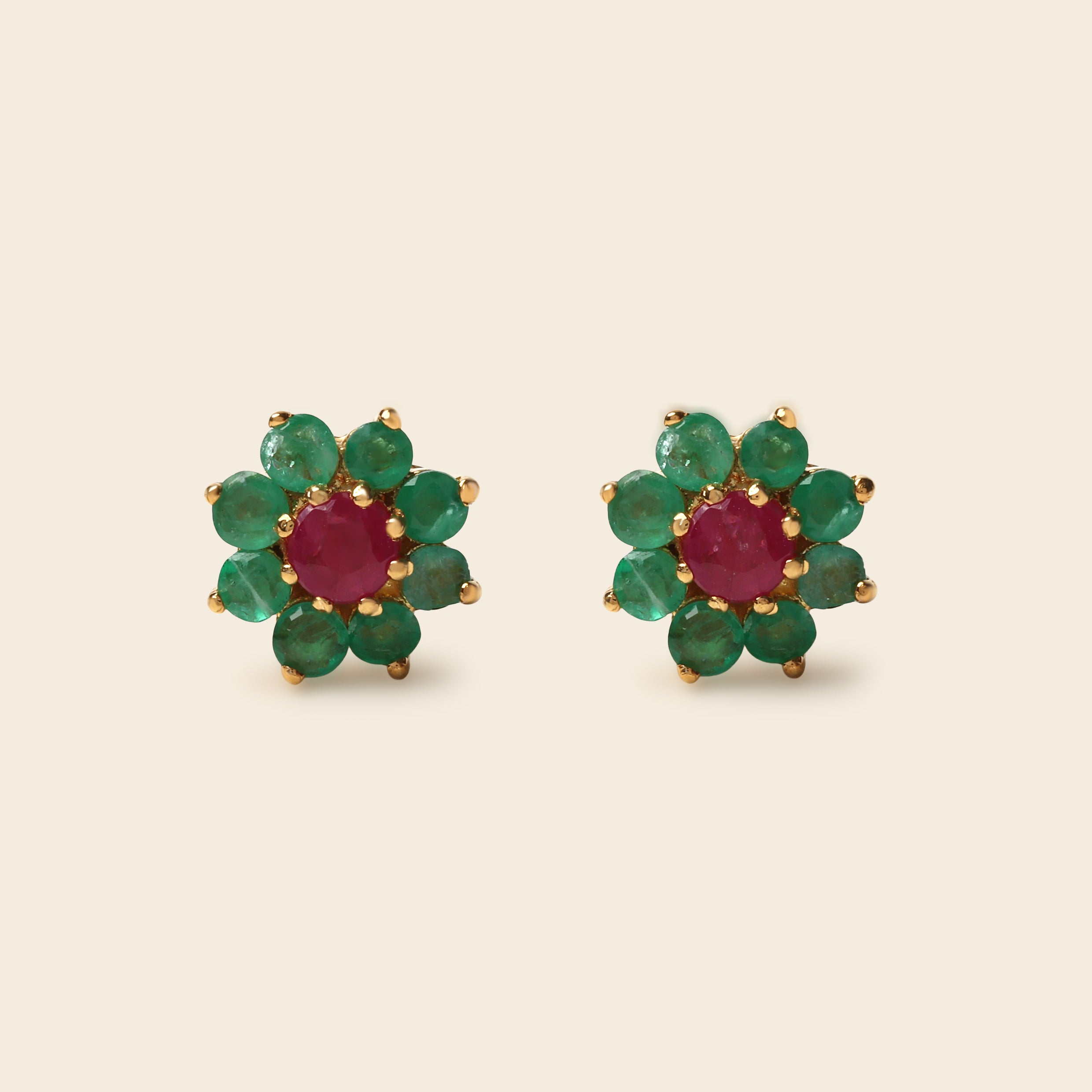 A pair of gold flower-shaped stud earrings featuring a round red center stone surrounded by six green round stones, set against a solid light beige background.