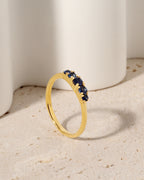 A gold ring with a row of dark blue gemstones, photographed on a textured beige surface with a soft-focus white background.