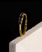 A yellow gold ring with a row of dark blue gemstones, displayed on a textured light beige stone block against a solid black background.