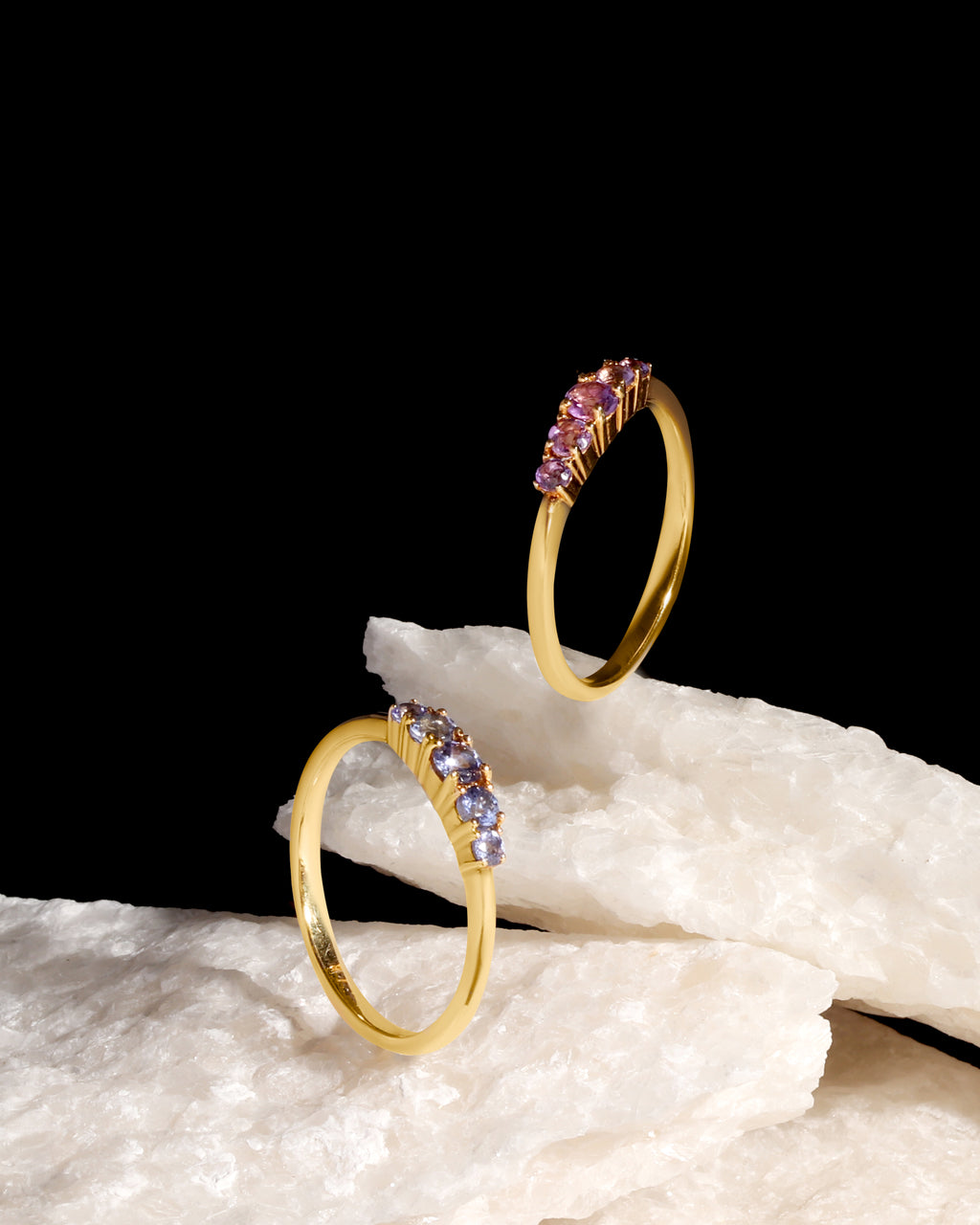 Two gold rings with small pink gemstone clusters, displayed on white textured stone against a solid black background.