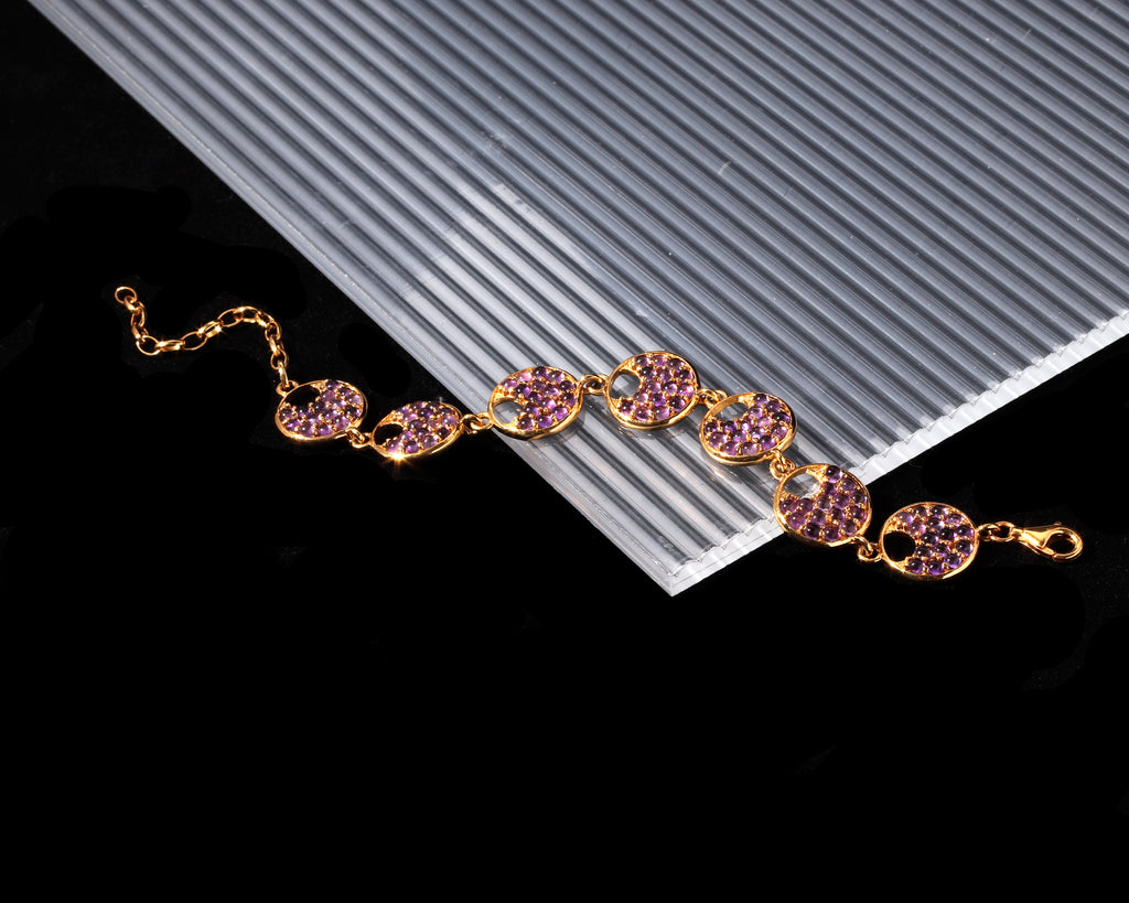 A gold-tone chain bracelet with oval links paved with pink gemstones, displayed diagonally on a white ribbed card over a black background.