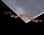 A gold-tone chain bracelet with oval links paved with pink gemstones, displayed diagonally on a white ribbed card over a black background.