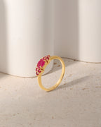 A gold ring with a marquise-cut pink gemstone and small round pink stones, displayed on a textured beige surface in front of white cylindrical props.