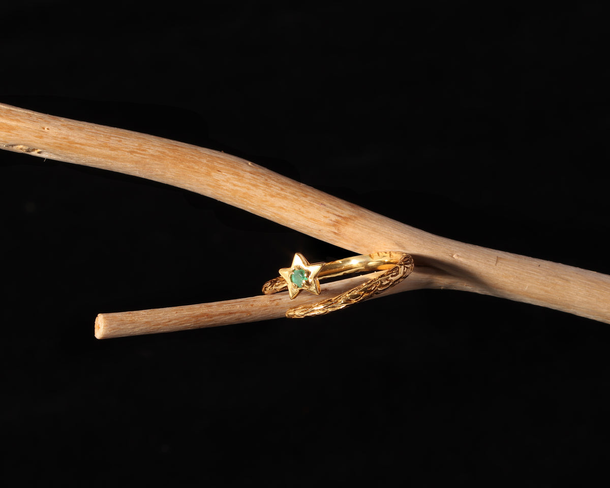 A gold ring with a star-shaped setting and a small green gemstone, displayed on a natural wooden branch against a solid black background.