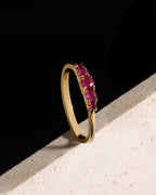 A gold band ring featuring a row of round-cut pinkish-red gemstones, photographed resting on a textured light beige surface against a solid black background.