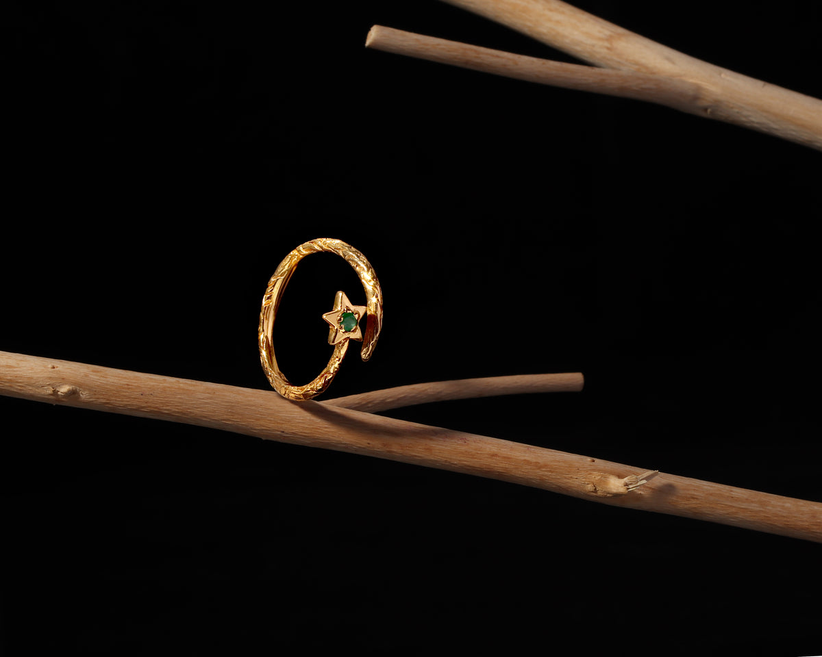 A gold ring with a textured circular band and a small star-shaped centerpiece set with a green gemstone, displayed on a natural wooden branch against a solid black background.
