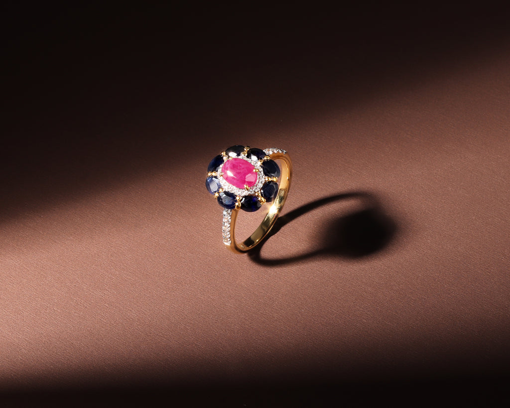 A gold ring with a central oval pink gemstone surrounded by dark blue round stones and small clear stones, photographed on a dark brown background with dramatic lighting and shadow.