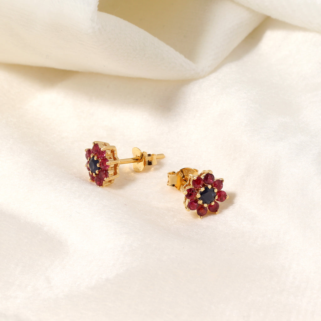 A pair of gold flower-shaped stud earrings with dark red gemstone petals and a dark blue center, displayed on a soft white fabric background.