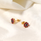 A pair of gold flower-shaped stud earrings with dark red gemstone petals and a dark blue center, displayed on a soft white fabric background.
