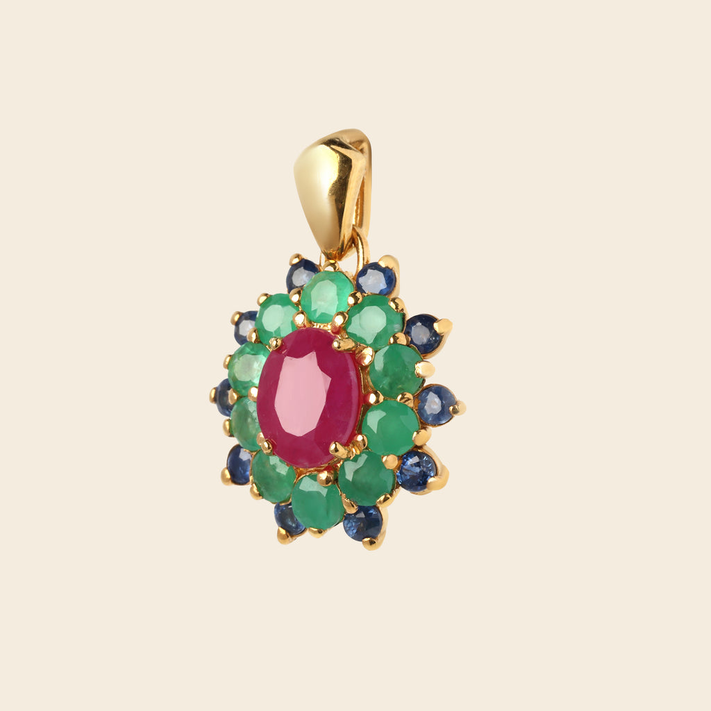 A gold pendant featuring a central oval pinkish-red gemstone surrounded by a double halo of round green and dark blue gemstones, set against a solid light beige background.