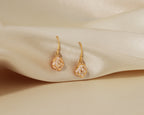 A pair of small gold rose-shaped drop earrings resting on draped cream satin fabric.