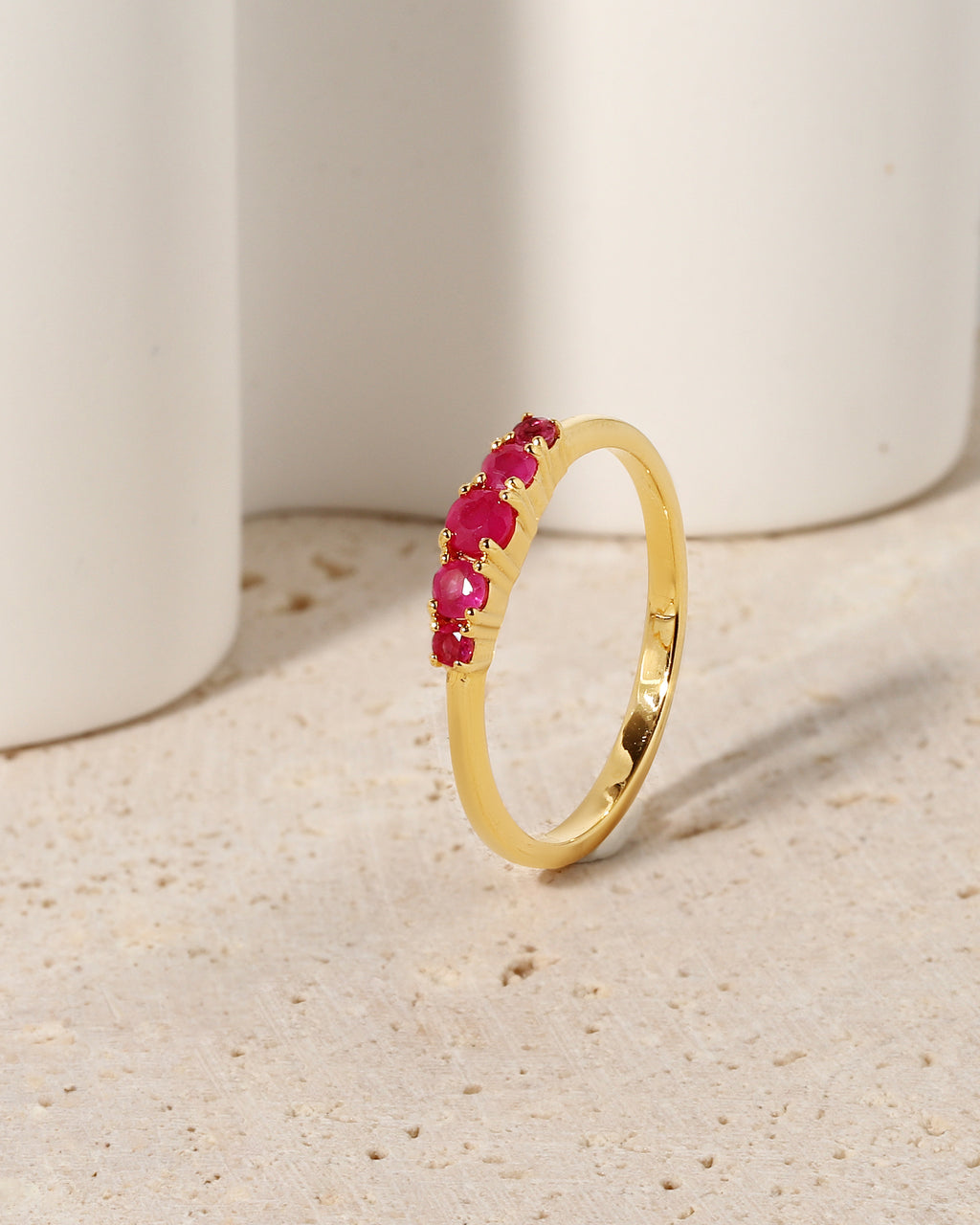A gold band ring featuring a row of small, round-cut pinkish-red gemstones, displayed on a textured beige surface with white ceramic cylinders in the background.