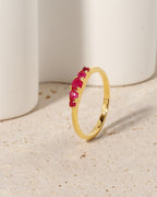 A gold band ring featuring a row of small, round-cut pinkish-red gemstones, displayed on a textured beige surface with white ceramic cylinders in the background.