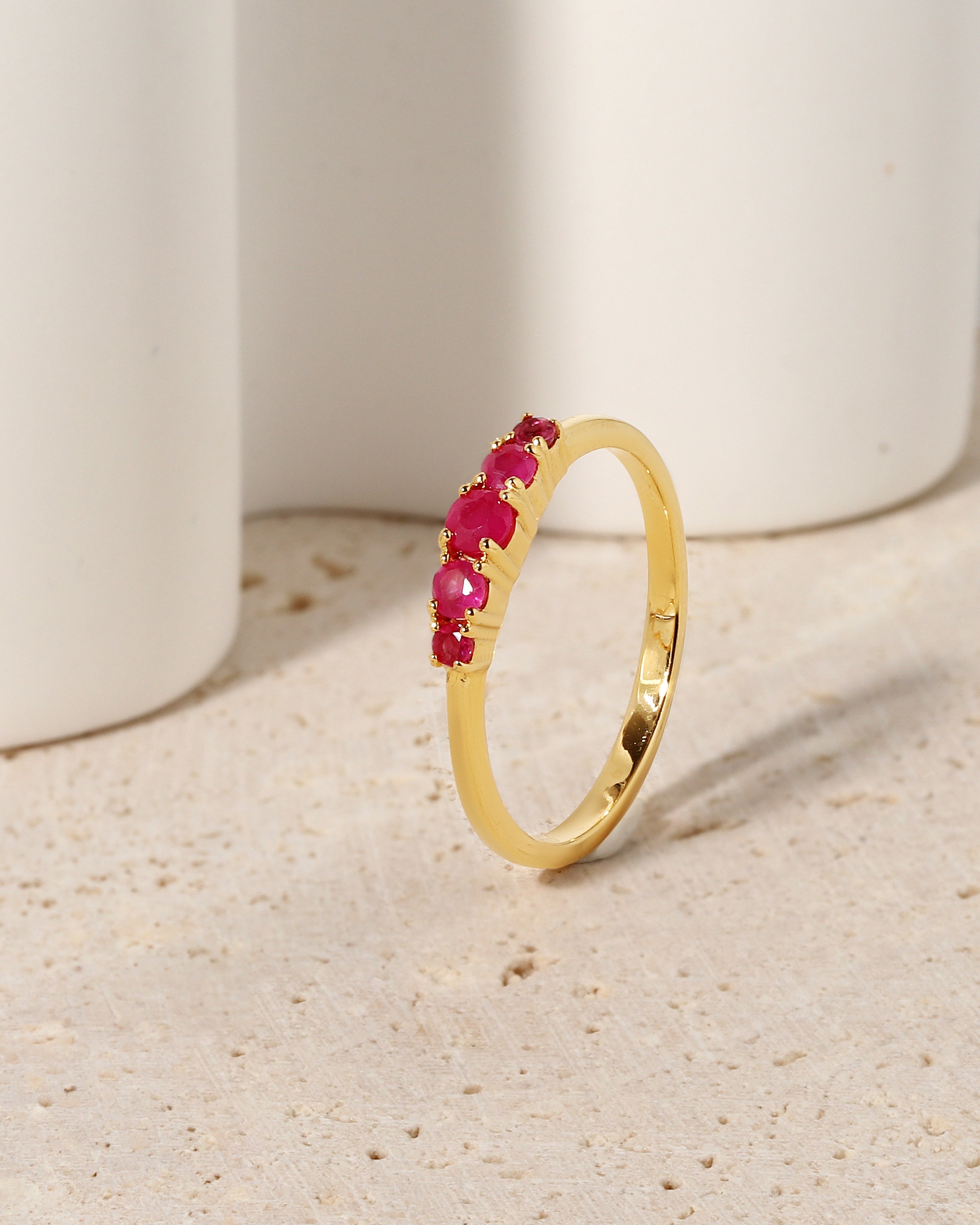 A gold band ring featuring a row of small, round-cut pinkish-red gemstones, displayed on a textured beige surface with white ceramic cylinders in the background.