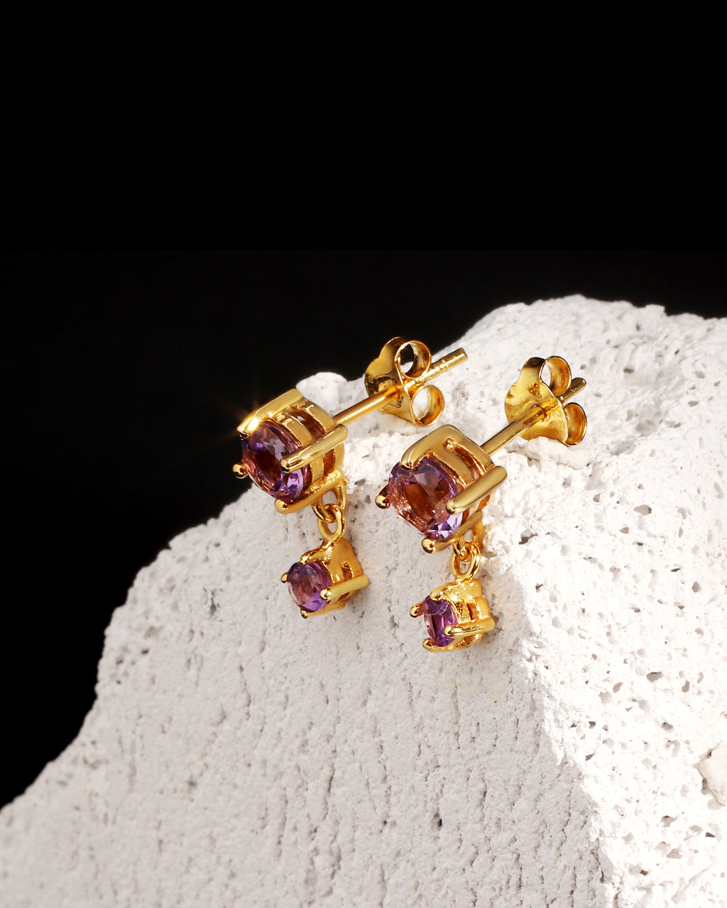 A pair of gold drop earrings with small square purple gemstones, displayed on a textured white stone surface against a black background.
