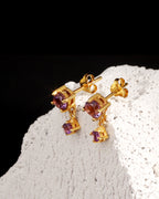 A pair of gold drop earrings with small square purple gemstones, displayed on a textured white stone surface against a black background.