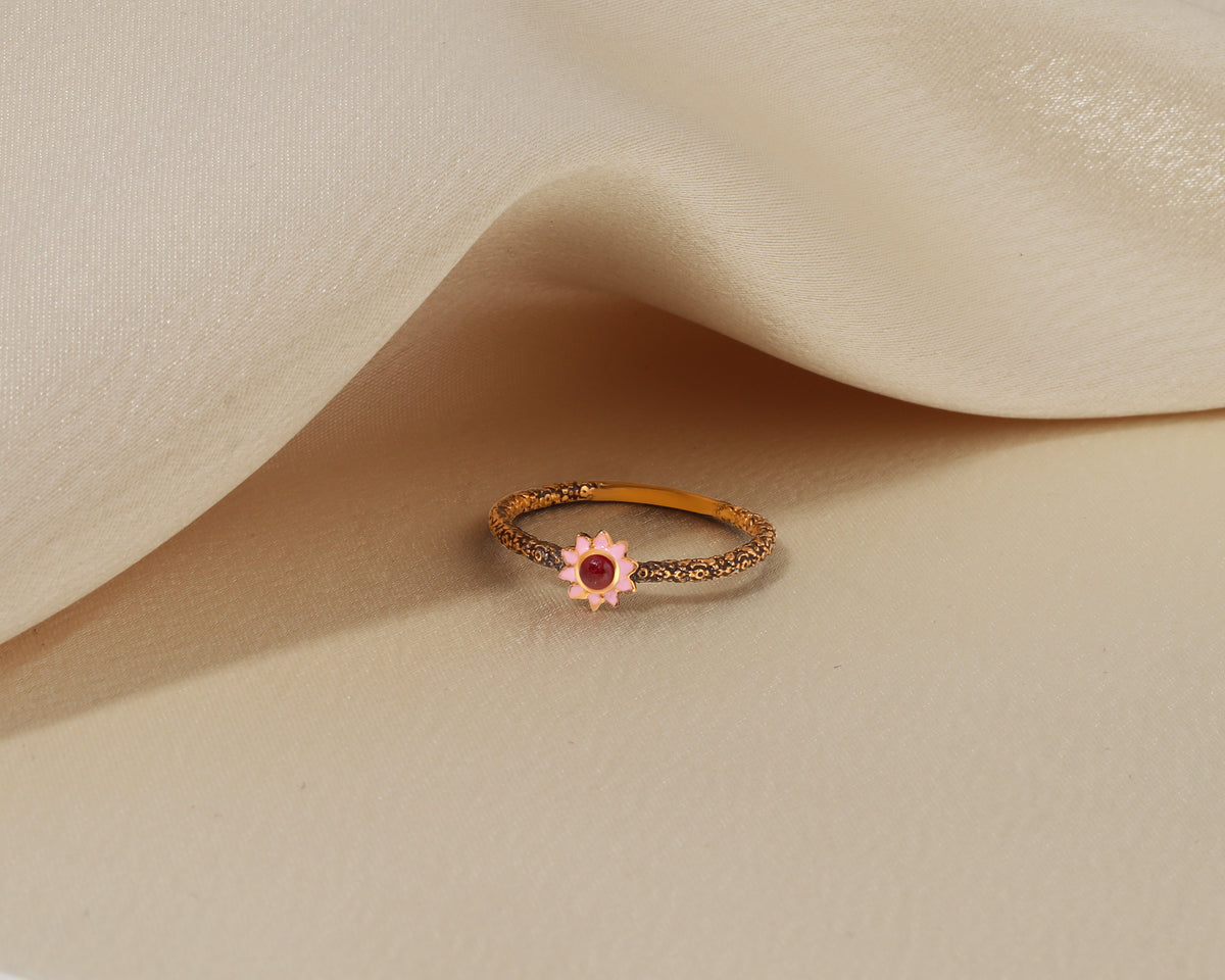 A delicate gold ring featuring a small pink flower setting with a central red gemstone, resting on draped beige fabric.