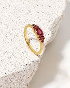 A gold ring with a oval-cut red gemstone and smaller round red gemstones, displayed on a textured white stone slab.