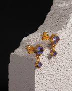 A pair of gold drop earrings with round blue gemstones, displayed on a textured white stone block against a solid black background.